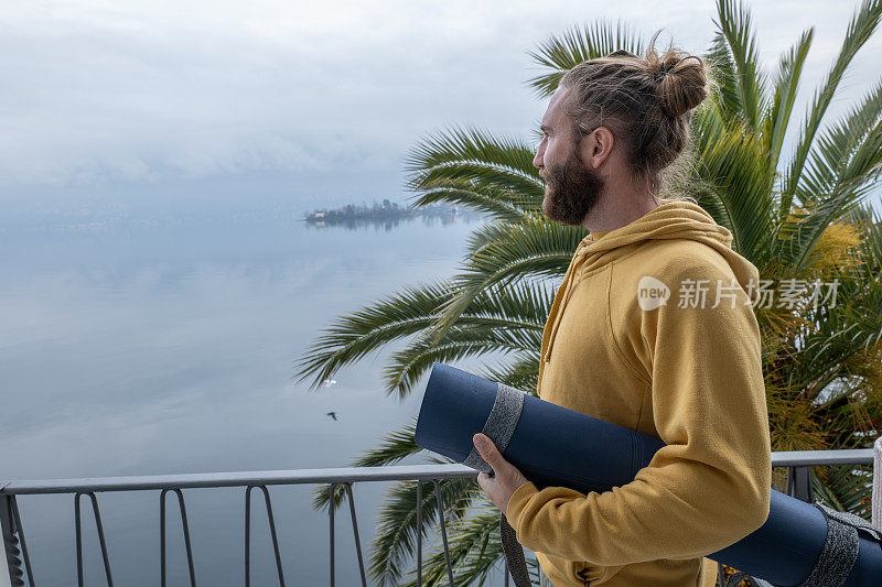 一个年轻人拿着瑜伽垫在她的阳台上沉思着湖和山的景色