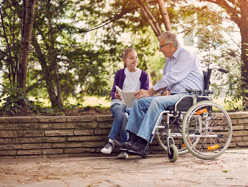残疾的爷爷坐在轮椅上在公园里和他的孙女一起看书