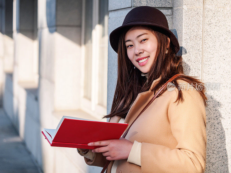 这是一幅美丽的中国女孩的肖像，她戴着棕色的帽子，拿着一本书，对着镜头微笑，背景是阳光明媚的建筑物。大学里的好读者。
