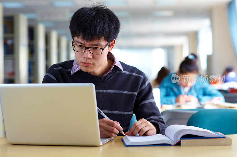 一名亚洲男大学生在图书馆学习，桌上放着笔记本电脑