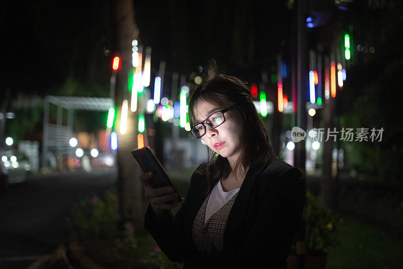 亚洲中国女人看着智能手机与担忧的表情脸在夜晚的街道上。