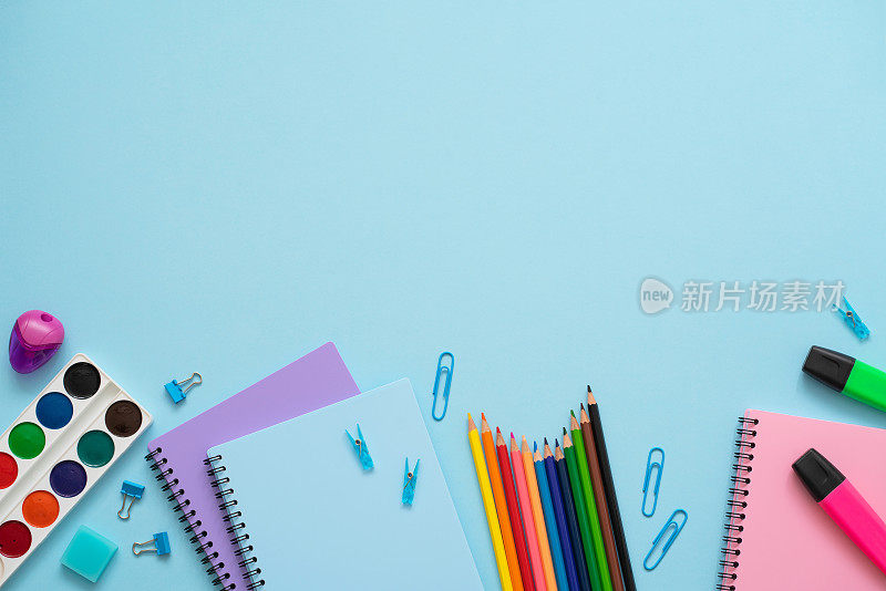 蓝色背景上是学校用品。顶部视图的蓝色，紫色和粉红色的笔记本，彩色铅笔，水彩画，回形针，马克笔和橡皮擦