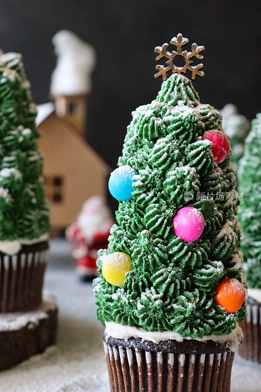 特写图像批量自制，圣诞树设计的巧克力纸杯蛋糕在纸杯蛋糕盒显示在雪，夜间针叶林的场景，模型房子与圣诞老人和棉花糖雪人