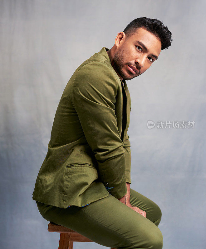 肖像的时装模特坐在凳子上与时尚，酷和时尚的绿色套装在一个工作室。迷人的印度男人坐在椅子上，穿着花哨，优雅和有品位的灰色背景的衣服。