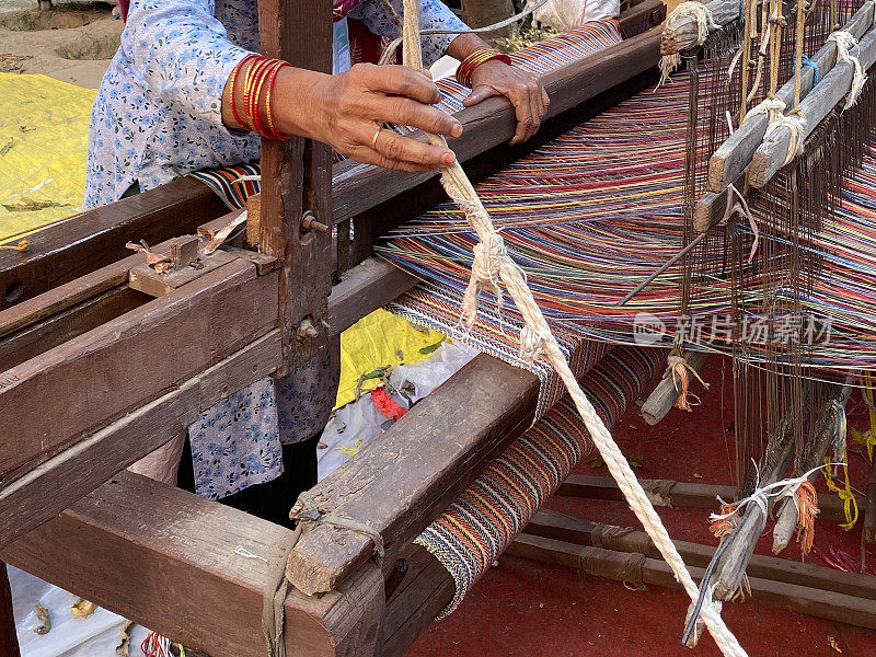 难以辨认的特写图像，印度妇女使用手工织布机编织彩色条纹地毯，经线，编结，踏板和布卷，重点在前景