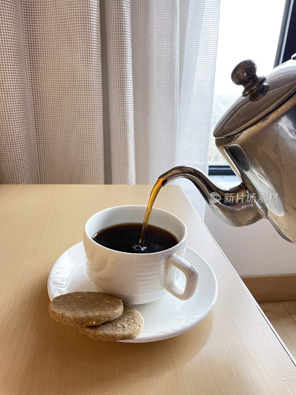 新鲜的黑咖啡从不锈钢咖啡壶的壶嘴中倒出来，白色的杯子和茶托配上两块饼干，酒店客房服务，重点在前景