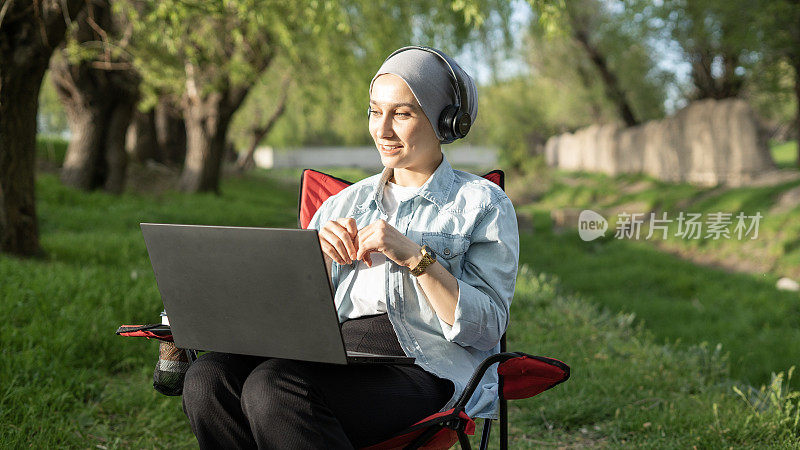 戴头巾微笑的年轻女子在自然公园用笔记本电脑视频聊天
