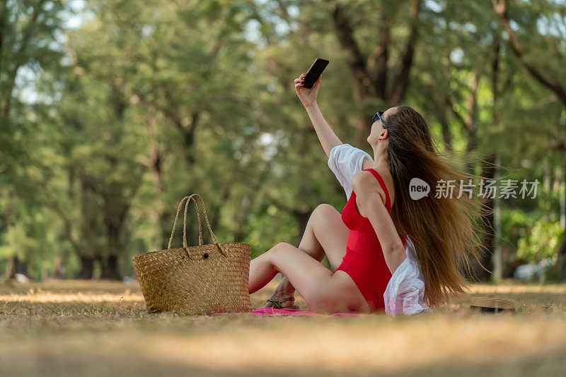 美丽的白人女性性感的女人在红色泳装比基尼戴着太阳镜自拍使用她的智能手机快乐和放松在海边的树林