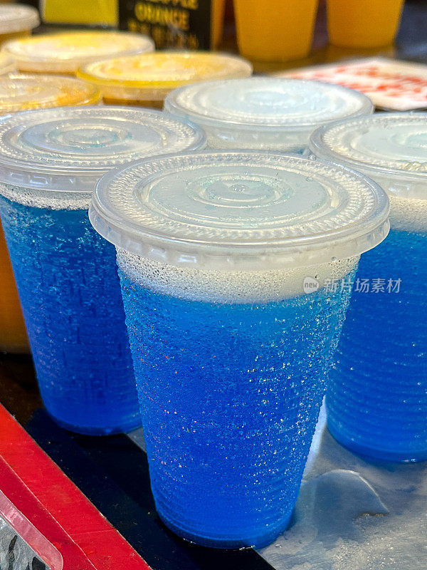 蓝色冰冻冰沙饮料装在有盖的塑料杯里，雪筒冰饮料装在一次性纸杯里，上面覆盖着凝结物，金属柜台上的人造彩色冰沙，这是前景的重点