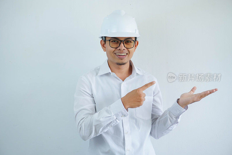 年轻的亚洲建筑师男子戴着白色安全帽，看着镜头，手指指向他张开的手掌，孤立在白色的背景上