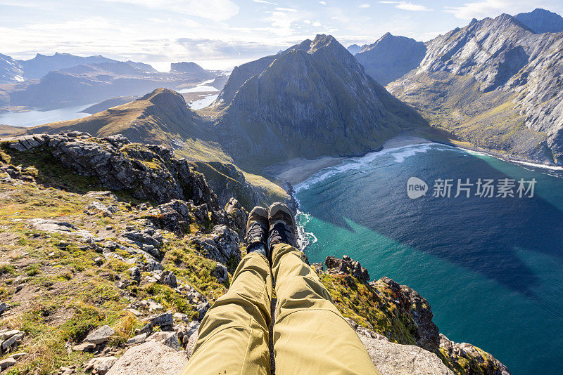 罗浮敦群岛上，一名男性徒步旅行者坐在一块悬空的岩石上，脚下是沙滩和山景