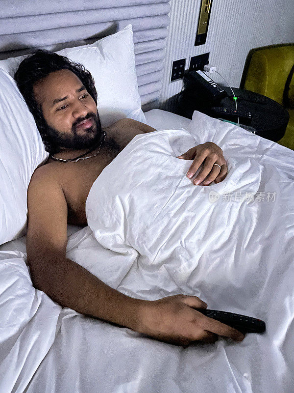 特写照片:印度男子躺在酒店的双人床上，躺在皱巴巴的床单上，靠着成堆的枕头，赤裸着胸膛，拿着遥控器换电视频道，抬高视野，聚焦于前景