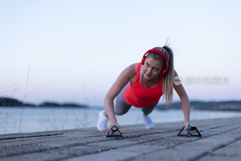 20多岁的金发美女在河边健身道边做俯卧撑边戴着红色耳机听音乐