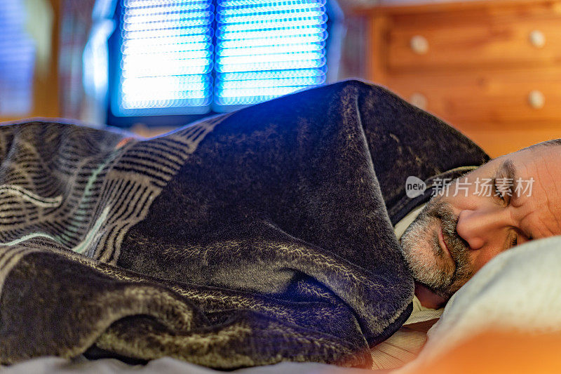 一个成熟的大胡子男人白天睡觉，躺在床上，盖着毯子，阳光透过百叶窗。