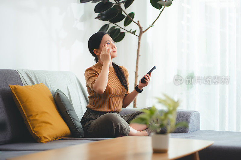 亚洲女性坐在家里的沙发上使用智能手机时会感到眼睛疼痛。