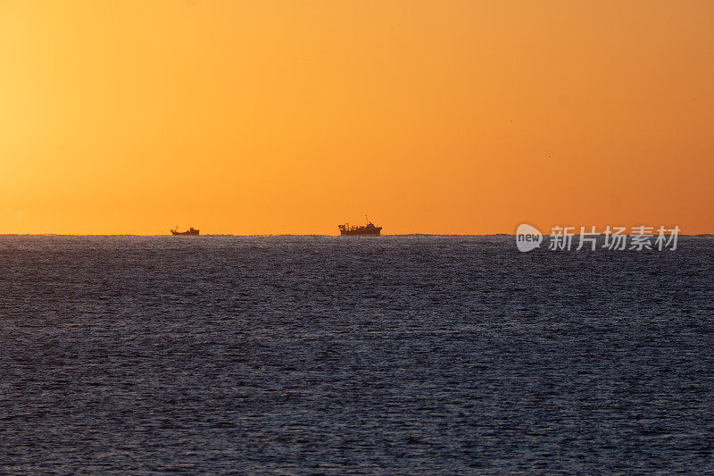 在橙色的天空下，地中海地平线上两艘渔船的剪影被黎明的太阳照亮，出海捕虾。