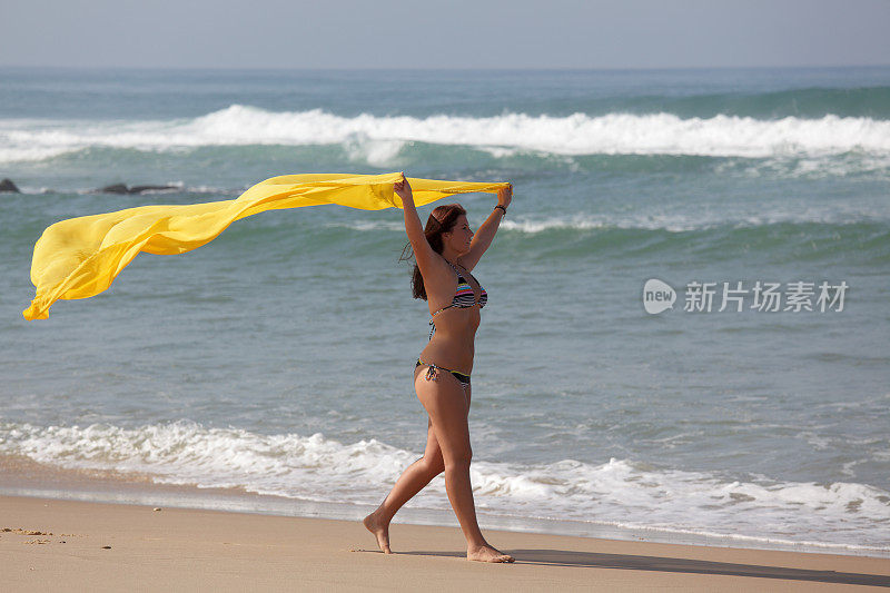 在风中拿着黄色围巾的女人走在沙滩上(XXXL)