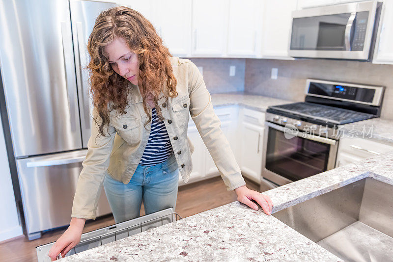 一名年轻女子在现代灰色、棕色的厨房里打开洗碗机，厨房里有花岗岩台面的橱柜和带有烤炉的瓷砖后挡板
