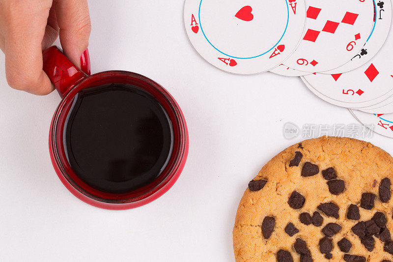 女手里拿着一个白色背景的红色咖啡杯，圆圆的扑克牌和一块饼干。