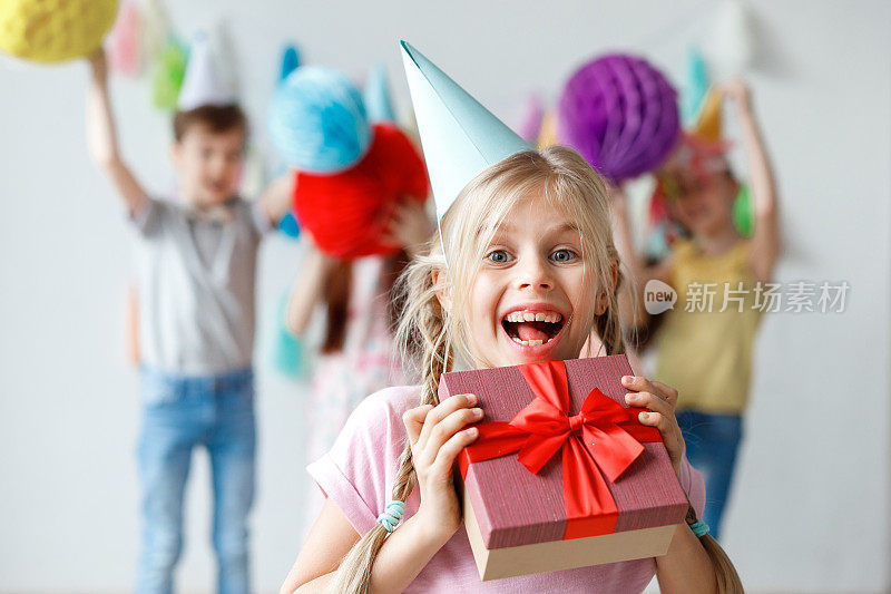 有趣的微笑美丽的小孩子戴着派对帽，拥抱大包装的盒子，高兴地收到来自亲戚的礼物，庆祝生日的朋友，小孩子们玩在背景。