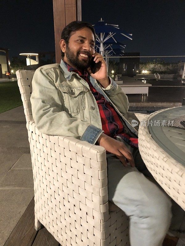 英俊的印度男子使用手机，坐在外面的咖啡桌在晚上捂着耳朵讲电话，笑着和微笑的手机照片，白色的柳条桌子和椅子庭院设置，玻璃顶部和黑色的夜空，家具