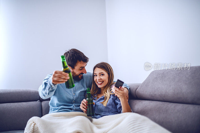 一对漂亮的白人夫妇盖着毯子坐在客厅里看电视喝啤酒。拿着遥控器换频道的女人。