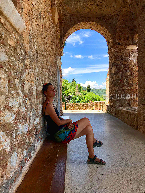 女游客坐在长凳下的历史拱廊与窗口像田园诗风景和蓝天在一个夏天的一天