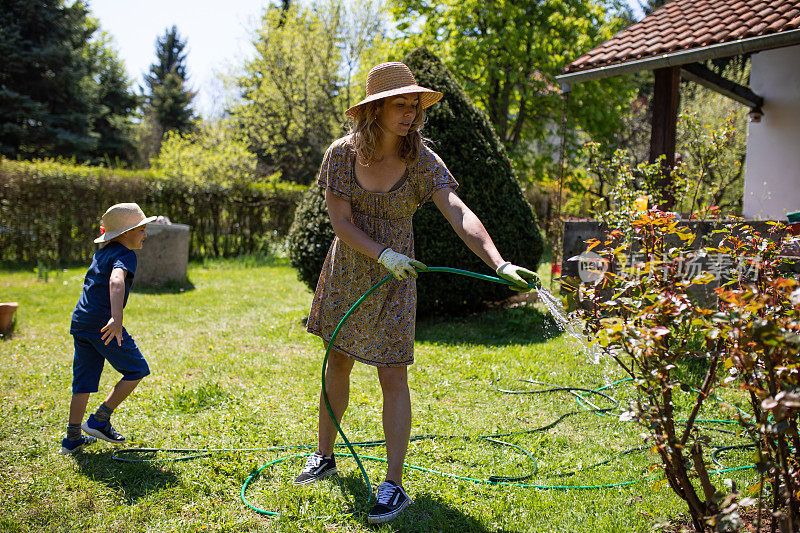 一个年轻的金发女人在给玫瑰浇水，她的儿子在他们的后院玩