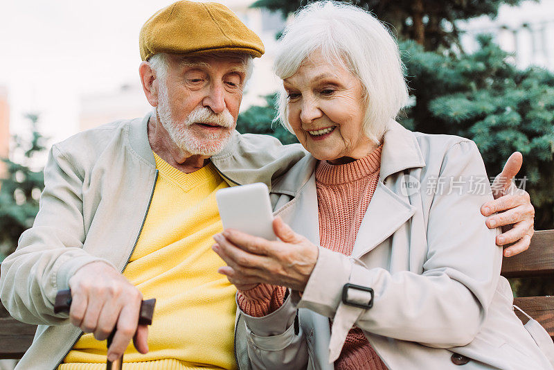 在公园长椅上，微笑的老妇人拿着智能手机靠近丈夫