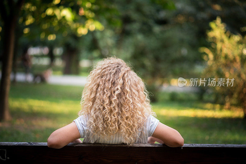 一位漂亮的卷发年轻女子坐在公园的长椅上，享受着阳光灿烂的日子