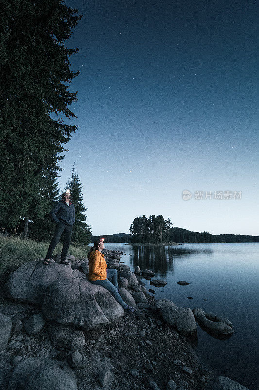 一起去摘星星。宽阔的镜头一对相爱的游客站在一块岩石在黄昏，看着雄伟的星空在一个美丽的山湖。灵感和灵性。心理健康。做梦