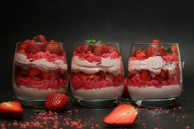 特写画面:三个杯子排成一排，里面有自制的草莓慕斯甜点，上面覆盖着整个草莓，奶油慕斯和红丝绒蛋糕屑分层布丁，周围是面包屑和水果，黑色背景，聚焦前景