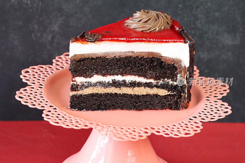巧克力和樱桃蛋糕片的特写图像，三个巧克力海绵层被奶油分开的粉红色蛋糕架，管道糖衣玫瑰花环，樱桃釉，斑驳的灰色和红色背景，重点在前景