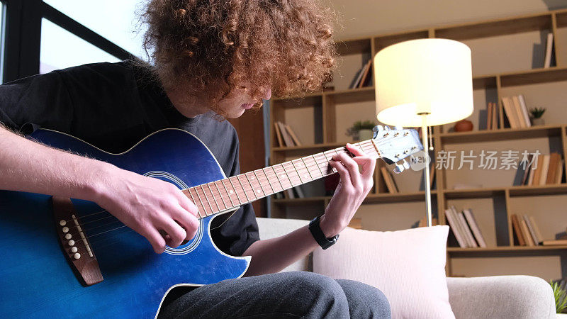 有创意的年轻卷发小伙音乐家坐在沙发上用原声吉他演奏旋律。