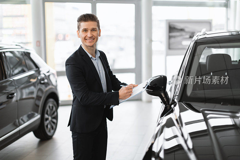 年轻的汽车销售员站在新车旁边，在汽车经销商复印空间里，对着相机微笑着做笔记
