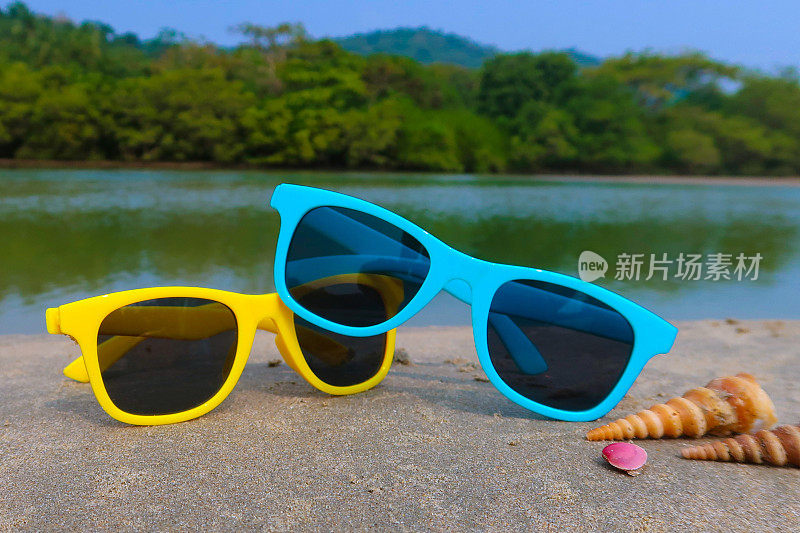 两副色彩鲜艳的儿童太阳镜，贝壳，蓝色和黄色的眼镜相互堆叠在印度海滩的岩石上，背景是大海和树木