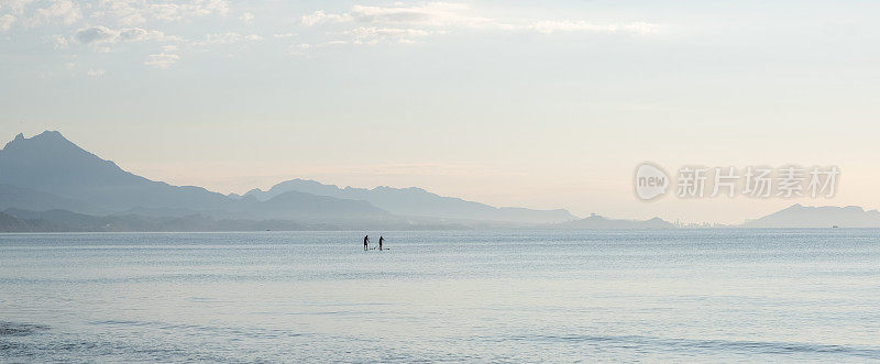 清晨的海景、山脉和海岸线。广阔的蓝色大海和背景中的两个人在充气冲浪板上。