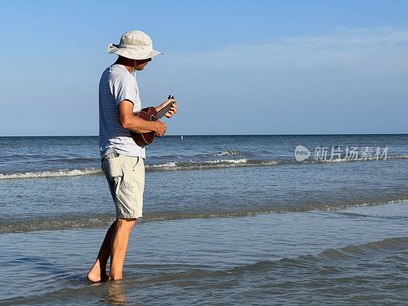 在佛罗里达海滩的浅水中赤脚行走的老人弹奏尤克里里琴的侧面视图。全长视图