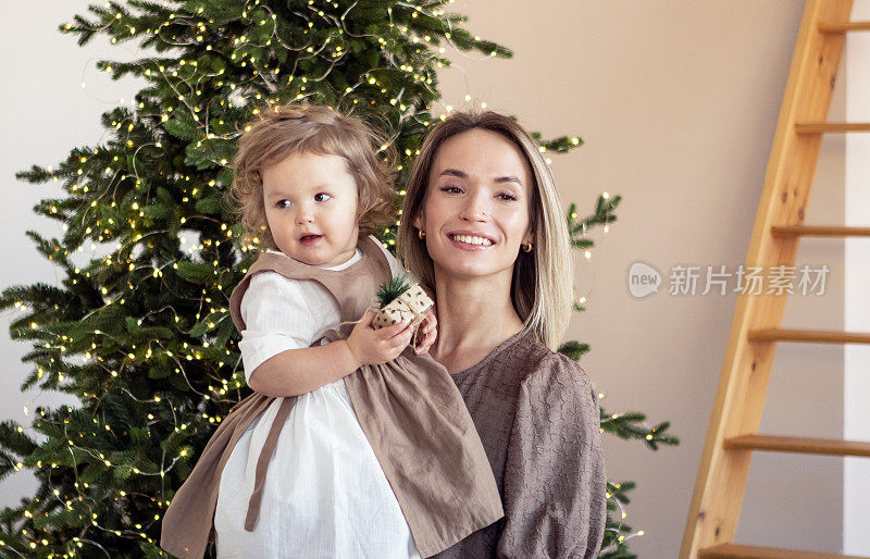 年轻的母亲和她金发碧眼的女儿在家里的圣诞树旁用礼物庆祝圣诞节。