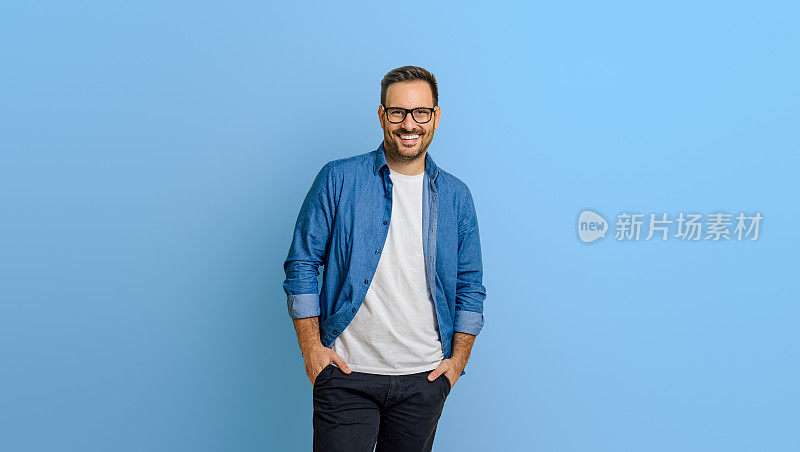 一个微笑的男性企业家的肖像，双手插在口袋里，自信地站在蓝色背景上