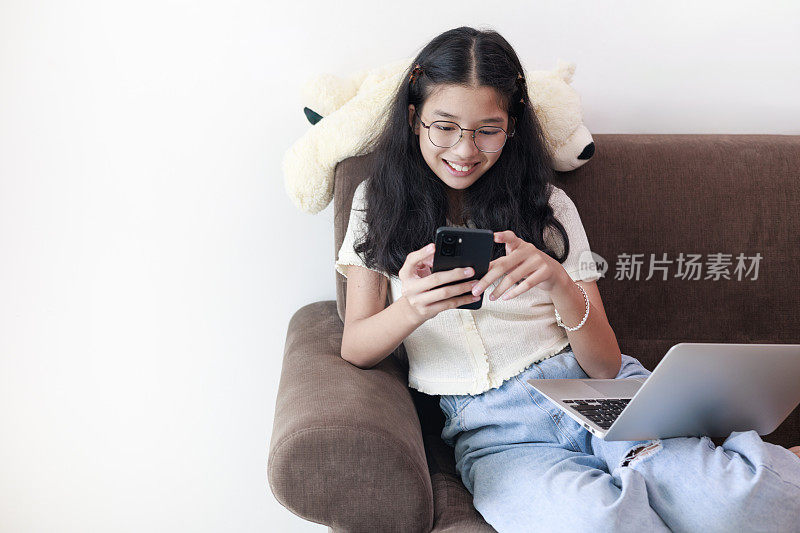 一个无忧无虑、放松的年轻亚洲女孩坐在沙发上玩她的智能手机。她正在浏览社交媒体、浏览互联网、网上购物或与朋友聊天。