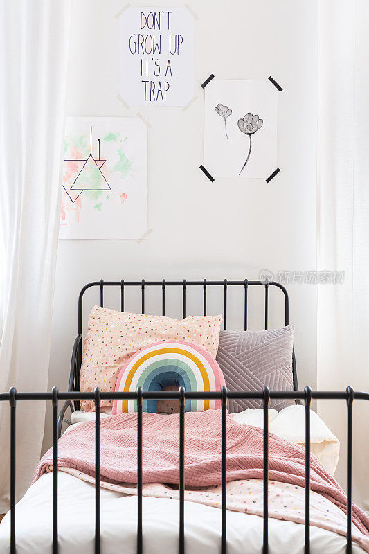 在白色的卧室里，孩子的床上有彩虹垫和粉红色的毯子。真实的照片