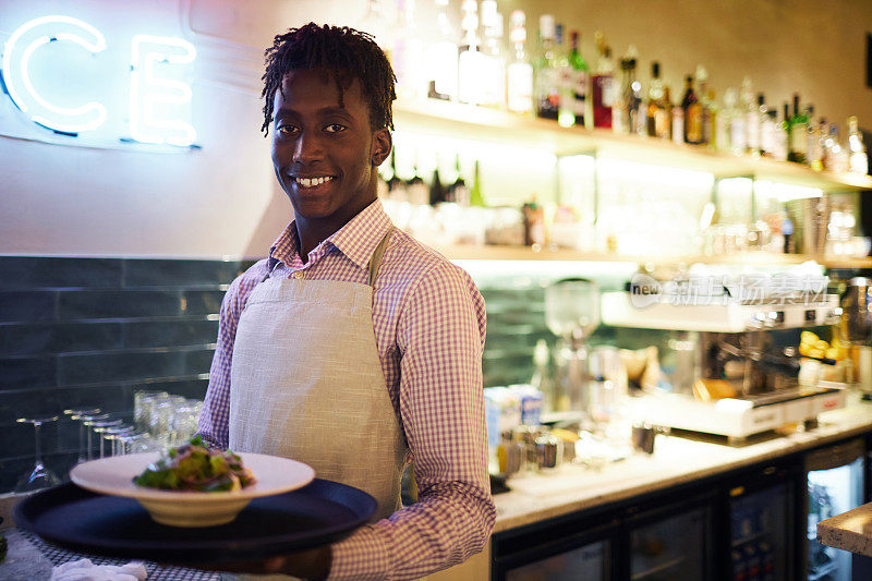 微笑自信的年轻非洲服务员，梳着辫，系着围裙，端着盘子，看着镜头在现代咖啡馆工作