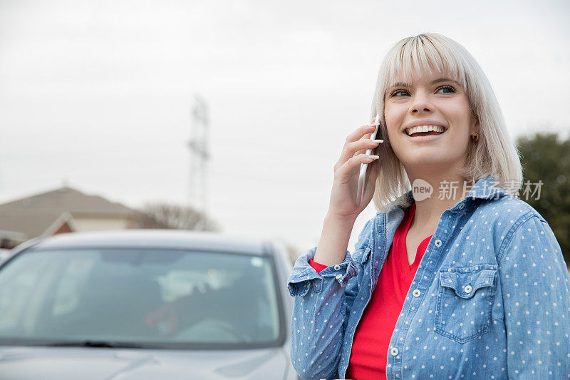 年轻的金发女人微笑着打电话，站在外面的车旁边