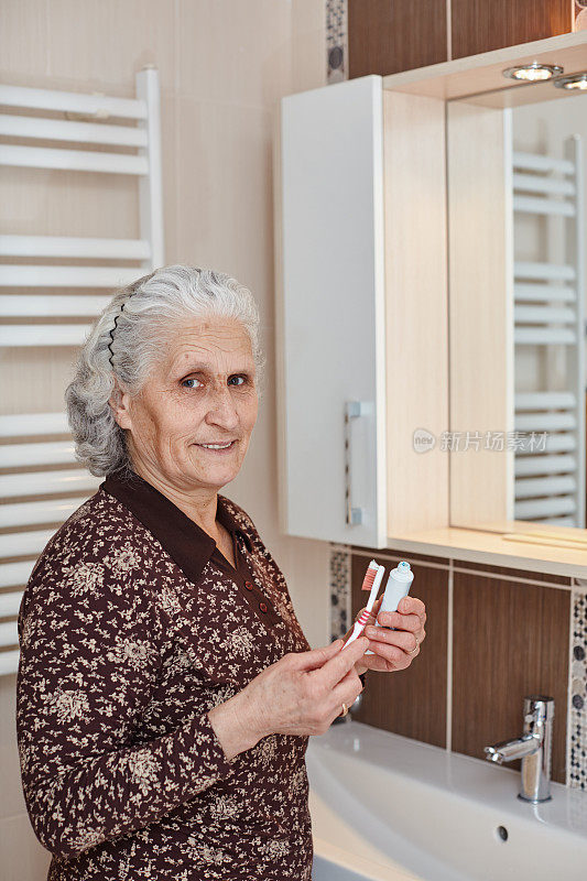 美丽的老妇人与灰色的头发拿着牙刷和牙膏在手里，而看着镜头前的浴室镜子和水槽