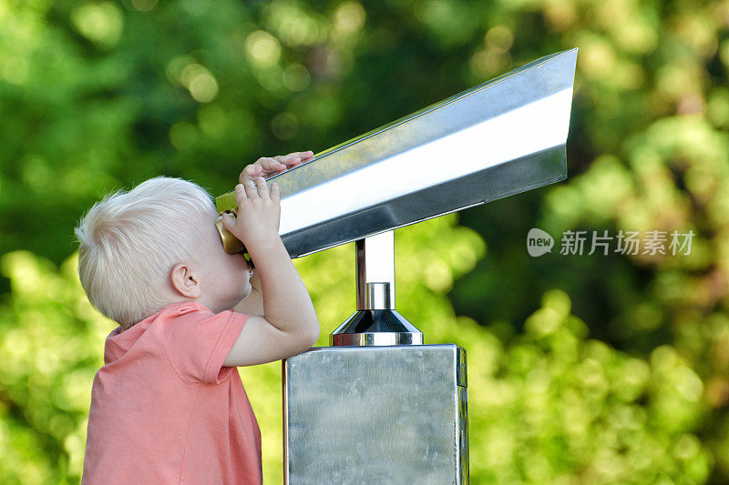 金发小男孩在看一副大望远镜。背景中的公园