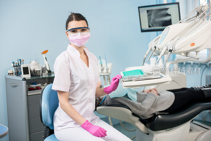 肖像有吸引力的女牙医与病人在牙科诊所。医生戴眼镜，口罩，制服和粉色手套。在背景监视器上用x光检查病人的牙齿。牙科