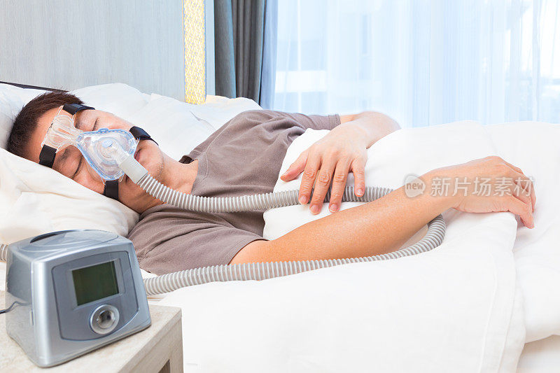 中年亚洲男子在床上睡觉，戴着连接呼吸软管和呼吸机的呼吸机面罩，呼吸机是治疗睡眠呼吸暂停的设备