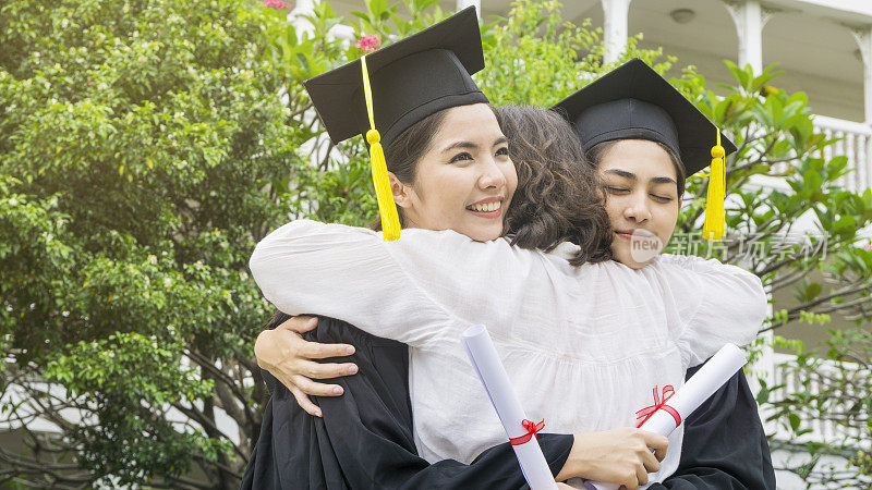两位亚洲女性身着毕业礼服，头戴毕业帽，微笑着与父母拥抱在一起