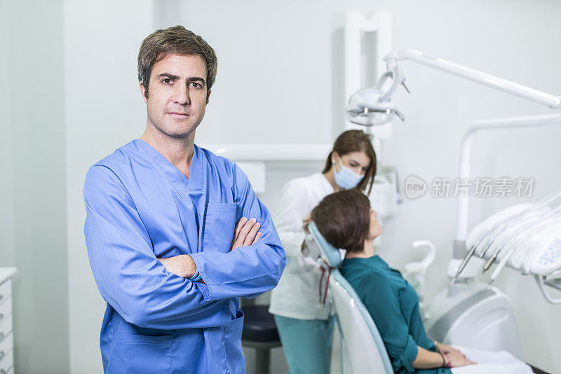 牙科诊所的牙医双臂交叉，看着镜头，病人和助手在后面